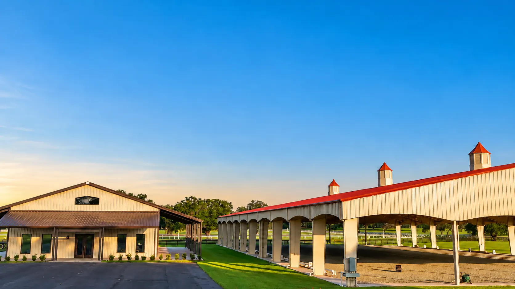 The long open-sided pavilion with its red roof and white cupolas at sunset, set beside the office barn and green grounds