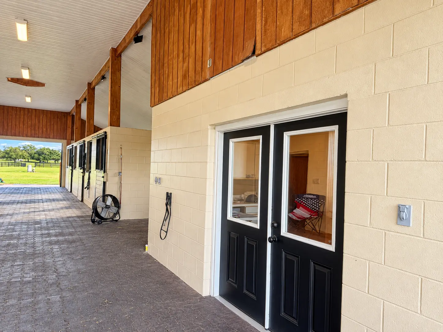 The barn aisle beside the office and tack room, its black glazed double doors set in a cream block wall beneath wood paneling, the aisle running to an open end onto sunlit pasture