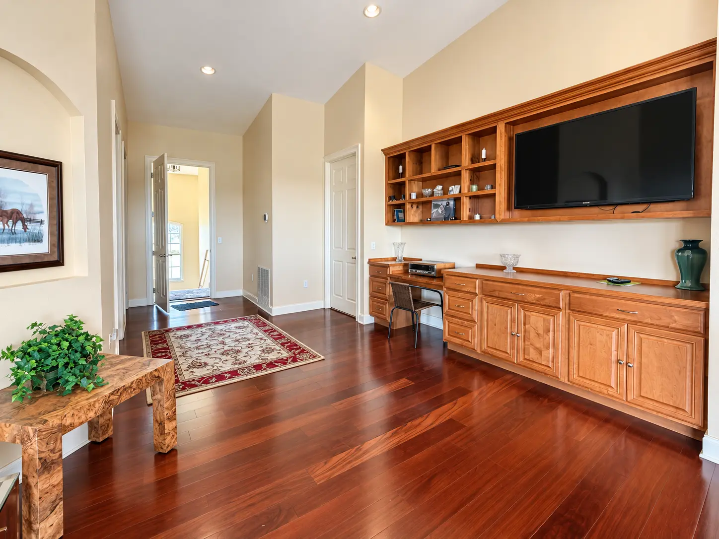 A sunlit sitting room in the mare-barn apartment: honey-toned built-in shelving and a long desk along one wall, glossy hardwood floors, an oriental rug in the hall, and a framed painting of a horse set in an arched niche