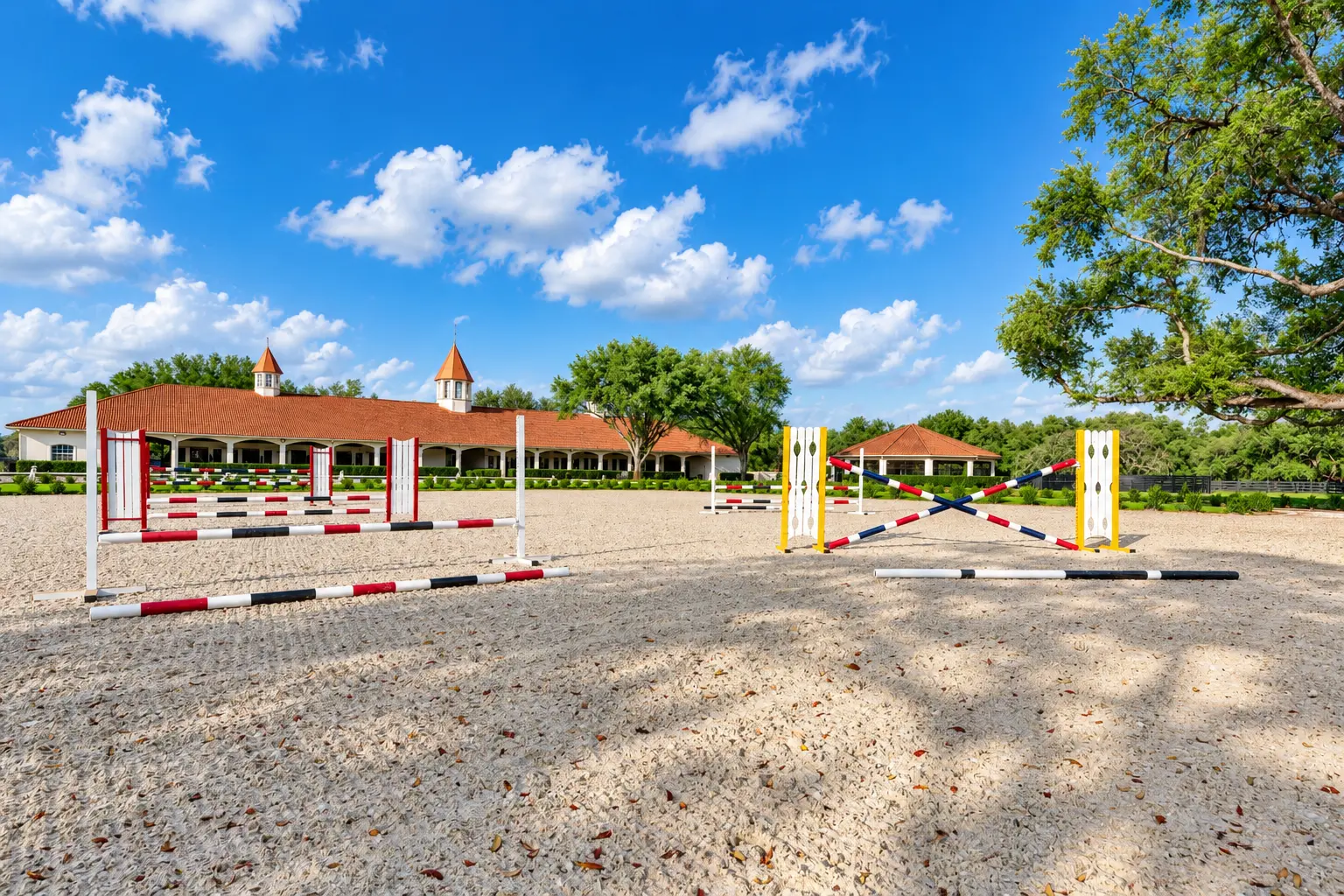 Red, white, yellow and blue jumping standards in the sand arena with the barn behind