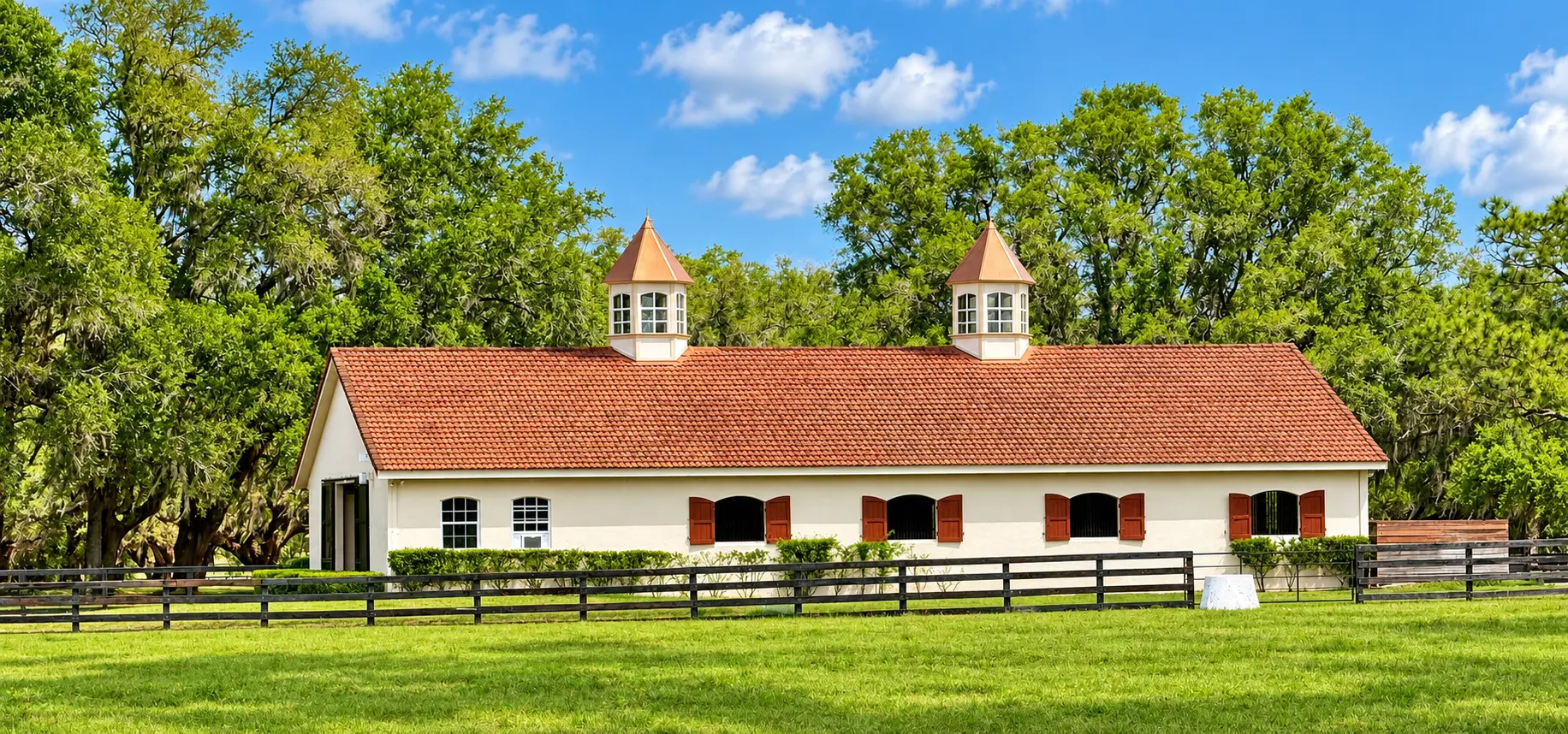 The Tudor Barn at GoldMark Farm — a long cream stucco barn with a terracotta tile roof, two cupolas and arched stall windows, fronted by a three-rail paddock fence beneath mature oaks