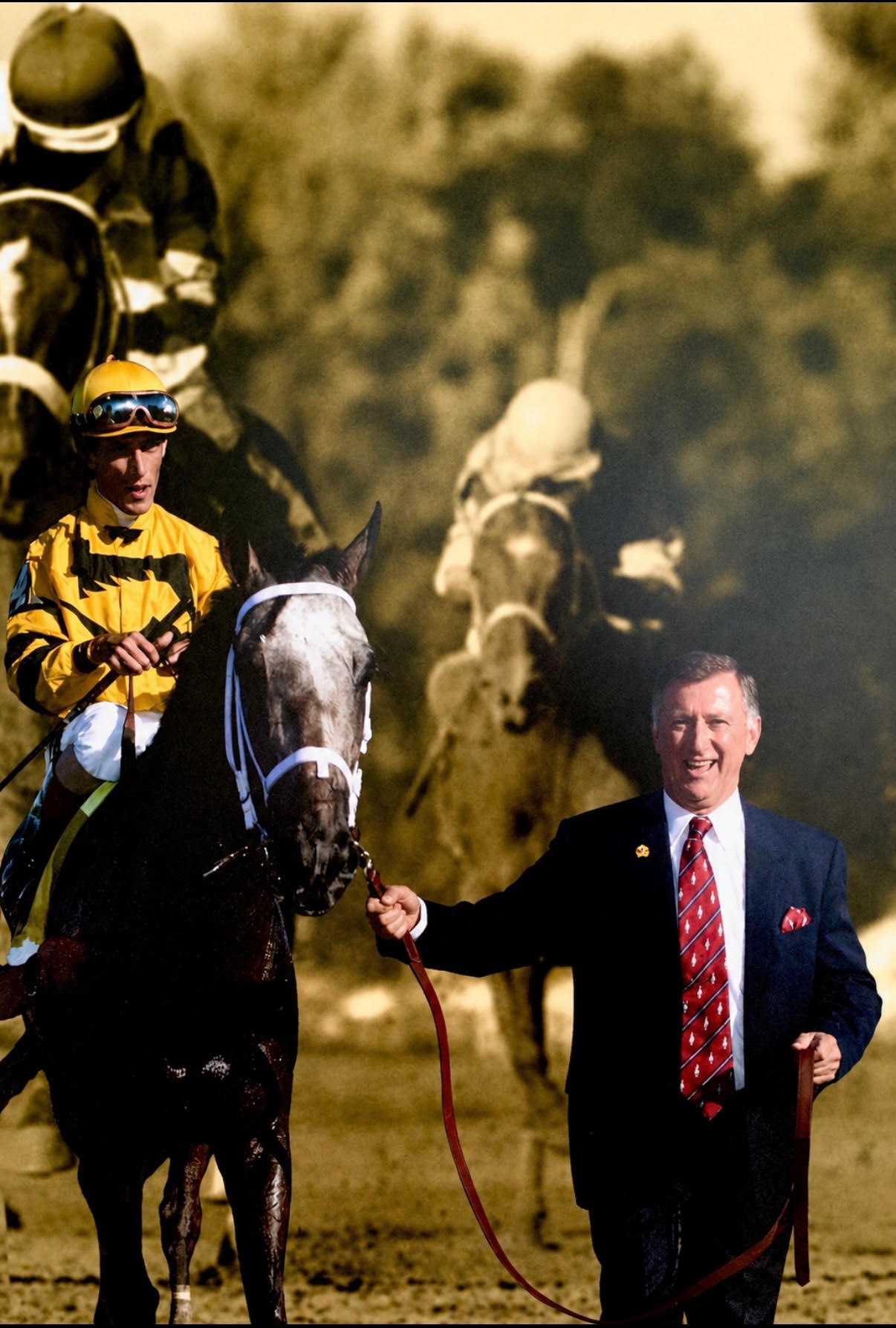 A jockey in yellow and black silks aboard a dark racehorse with a white blaze, led along the track by the founder in a navy suit and red tie, with racehorses running in the background