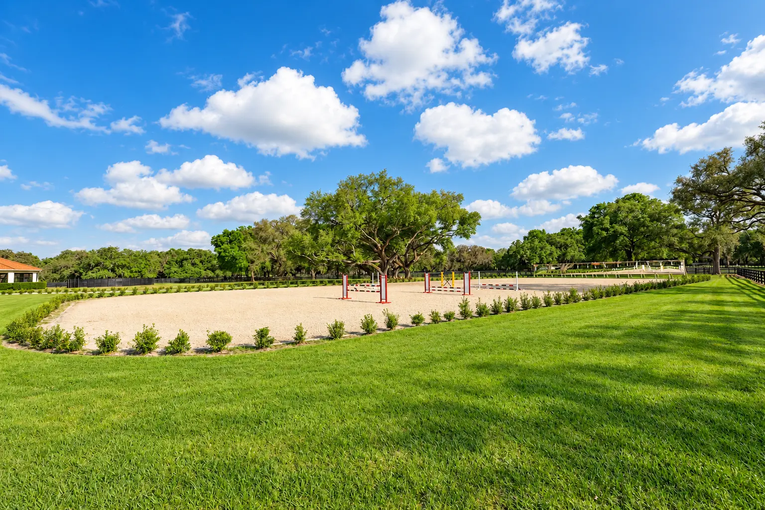 The sand jump arena set with jumping obstacles, bordered by hedges, lawn and mature trees