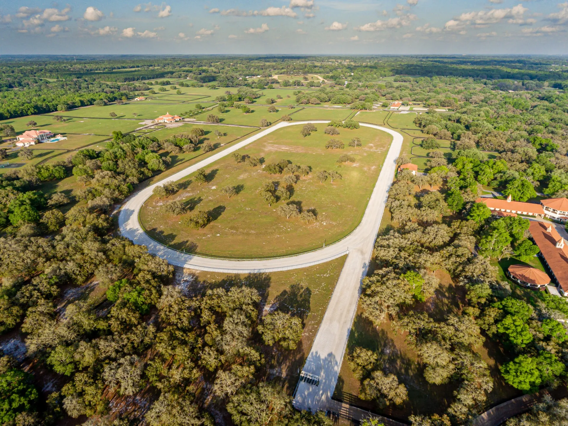 Aerial photo of the GoldMark ¾-mile training track — the sand oval looping between moss-draped oaks and green paddocks with the red-roofed barns and office complex beyond