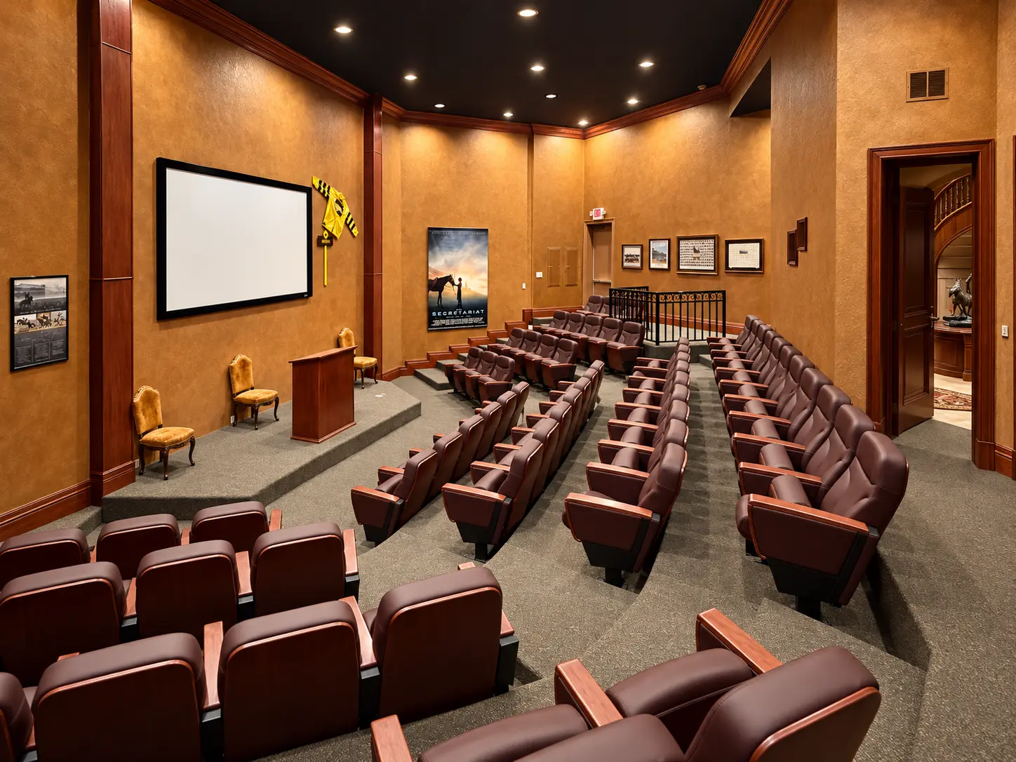 The estate theater in warm light: tiered rows of dark leather seats facing a small stage with a lectern, a projection screen at the side and equestrian prints along the walls