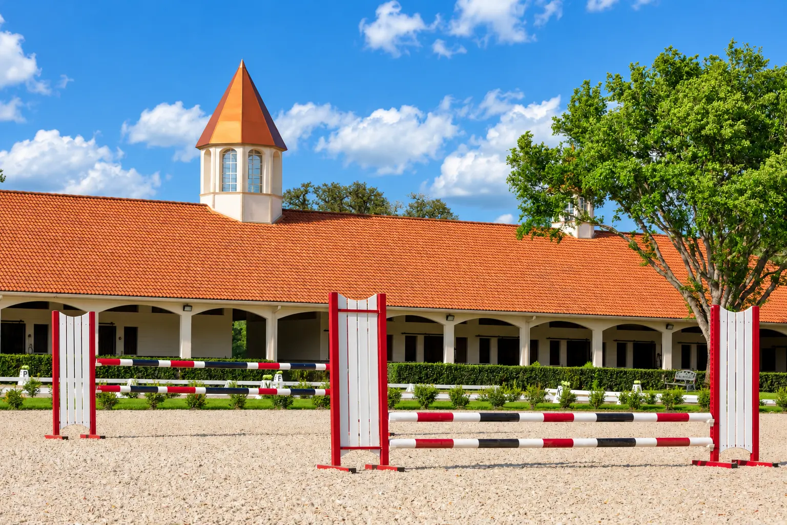 Colorful show-jumping obstacles set in a sand arena with a terracotta-roofed stable building and cupola behind