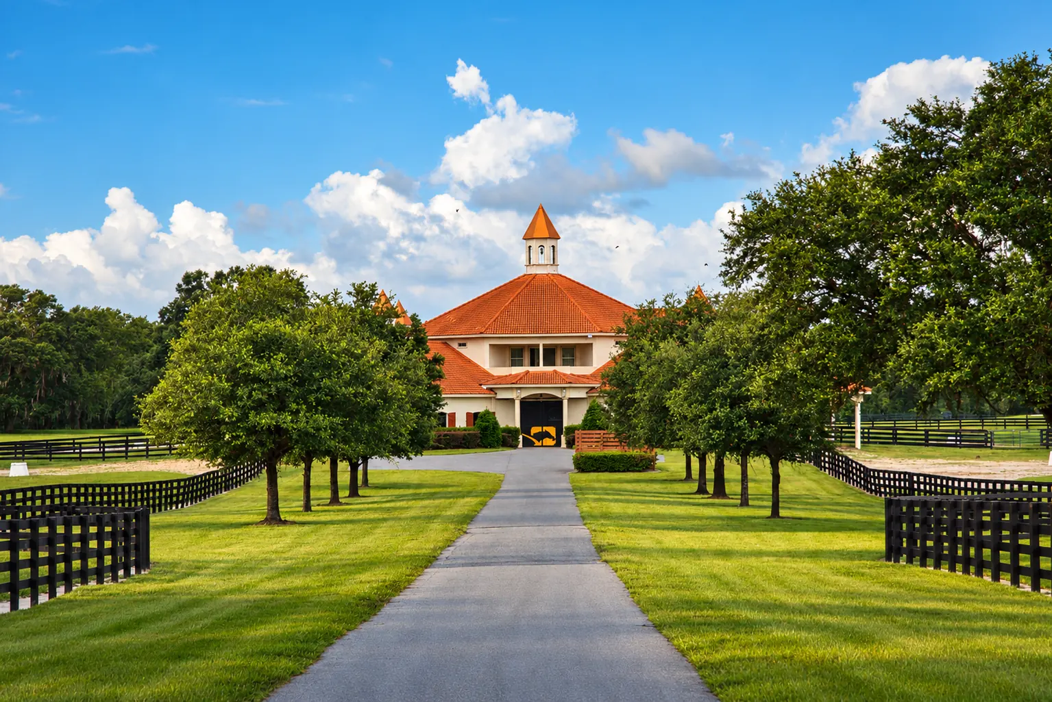 The CEM quarantine barn at GoldMark Farm — a cream stucco barn with a terracotta tile roof and central cupola, approached along a paved driveway lined with black post-and-rail fencing and mature trees