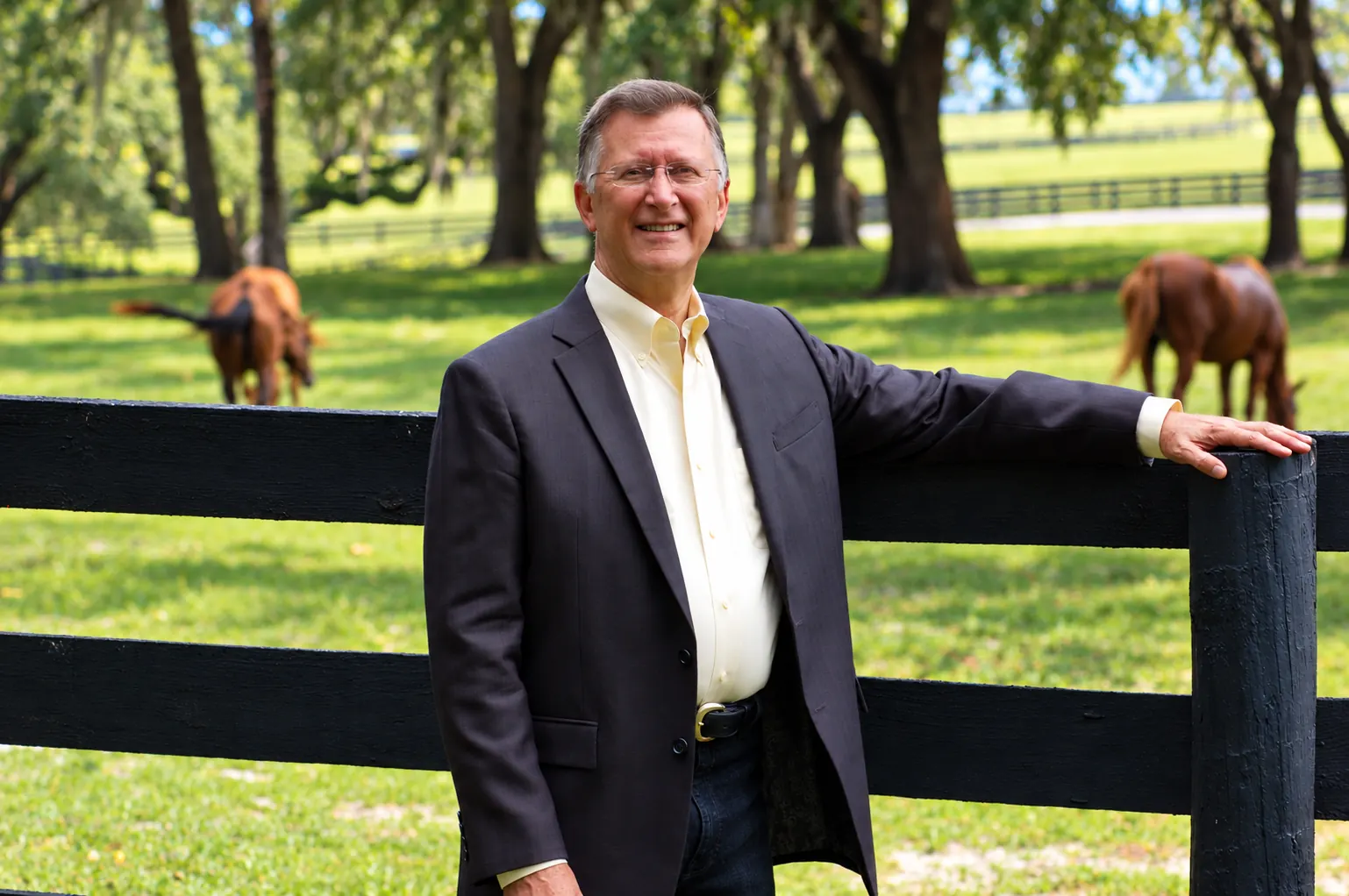 The founder in a suit jacket resting his hand on a black pasture fence, two horses grazing beneath the oaks behind him