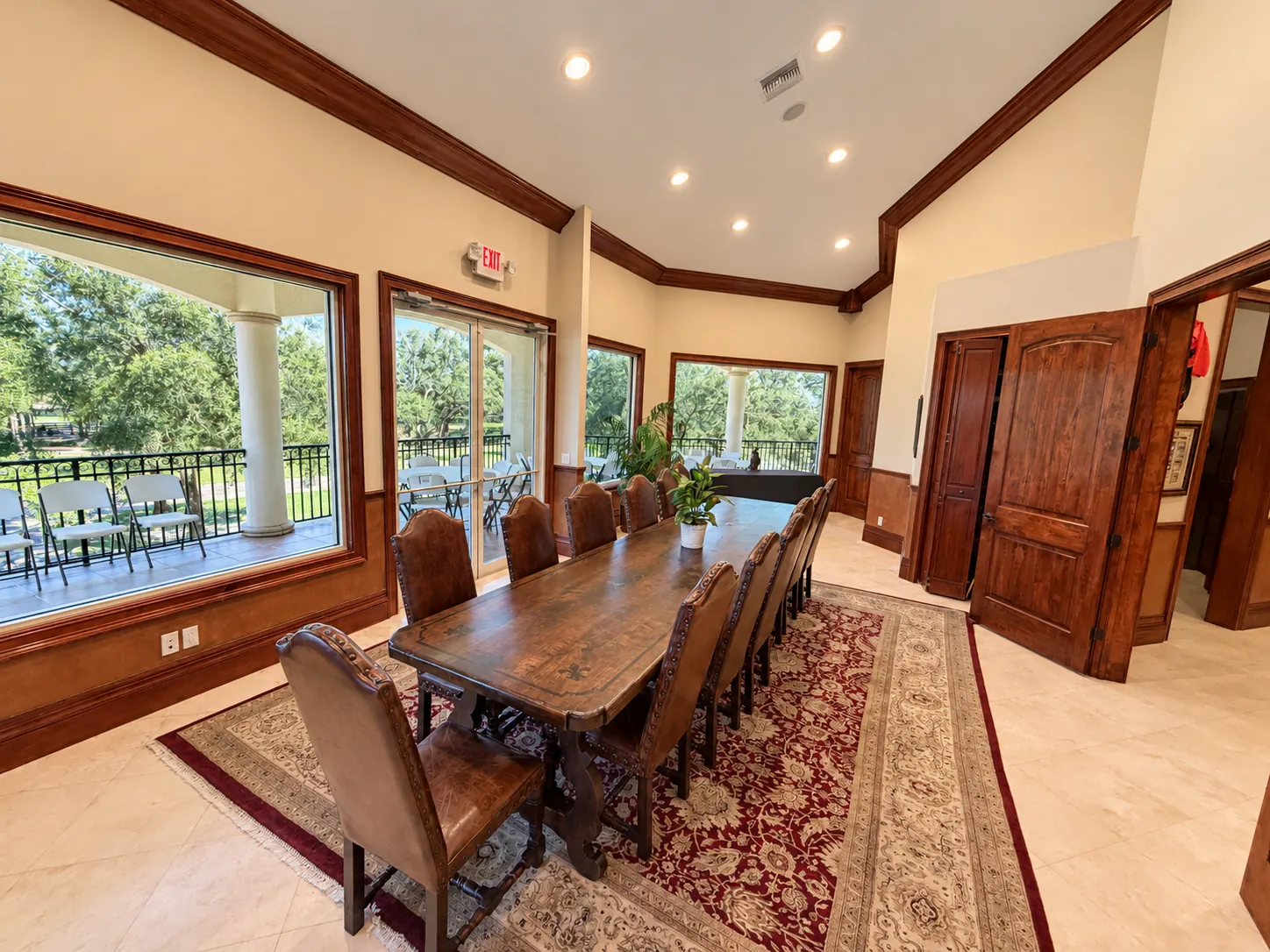 A board room in warm wood: a long dark-wood table ringed by high-back leather chairs on an oriental rug, glass doors opening onto a covered porch with round columns and green lawns beyond