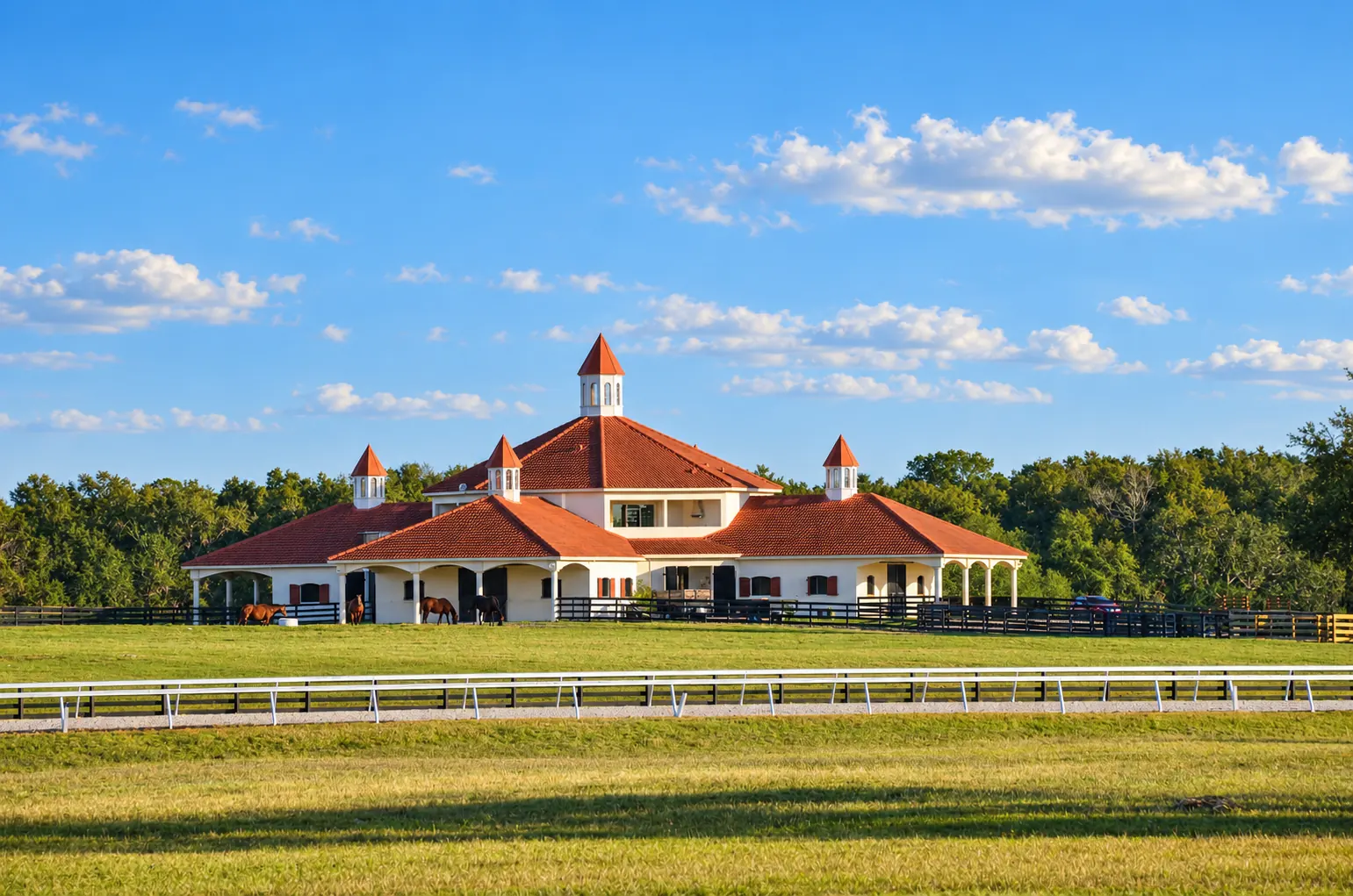 The mare foaling barn at GoldMark Farm in golden light — a long cream stucco stable complex with terracotta tile roofs, arcaded walkways and white cupolas, horses grazing in the paddocks out front and the track running along the fence line