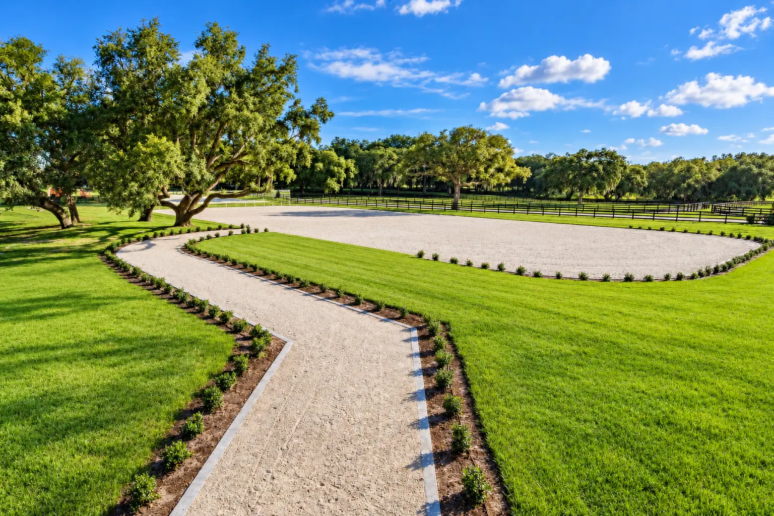 An outdoor sand riding arena joined to a winding sand path bordered in stone beneath mature oaks