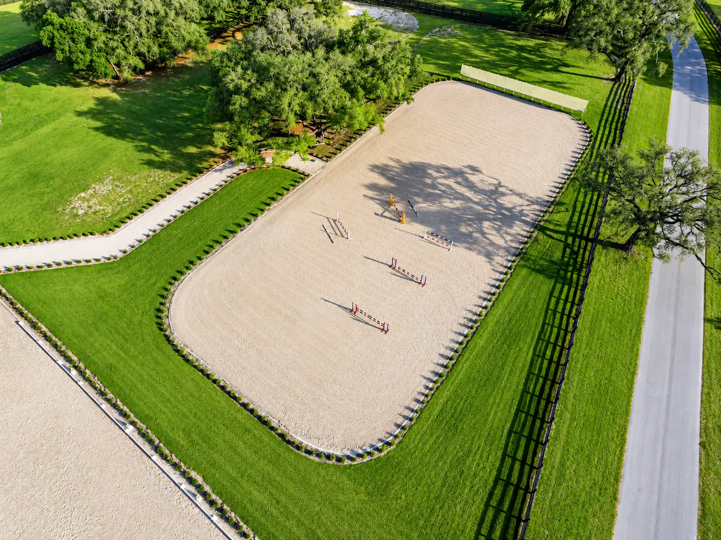 Overhead aerial of the sand jumping arena with a tree shadow across the footing and paddock fencing beyond