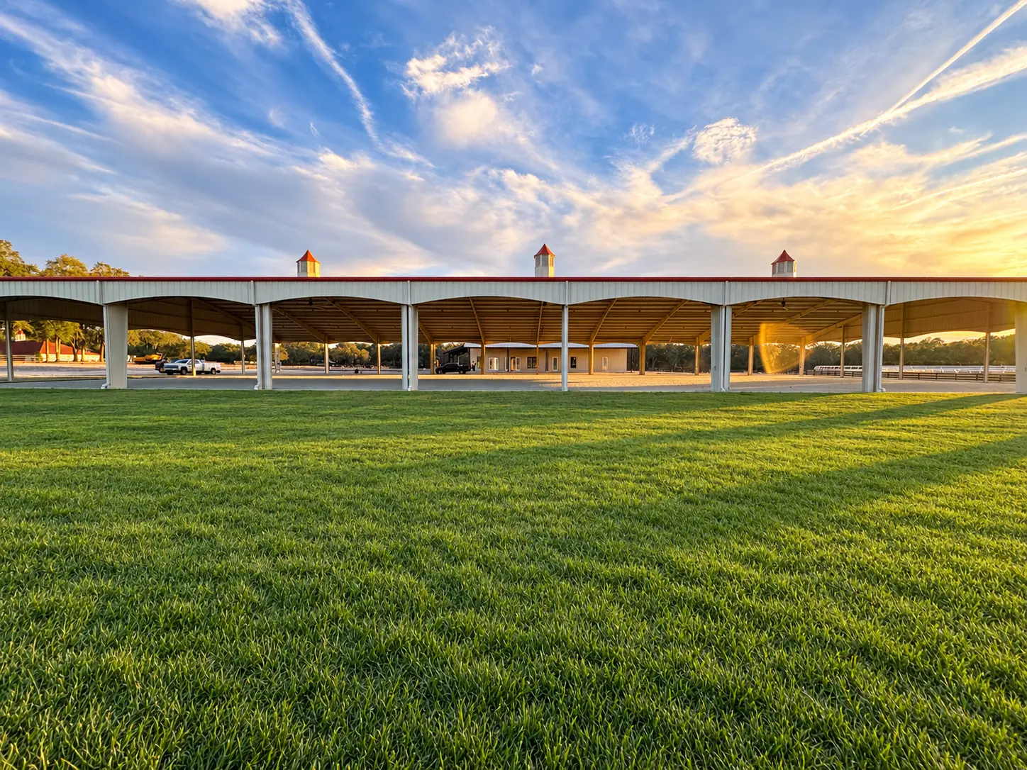 The covered arena's long roofline with its red trim seen across a lush green lawn at sunset
