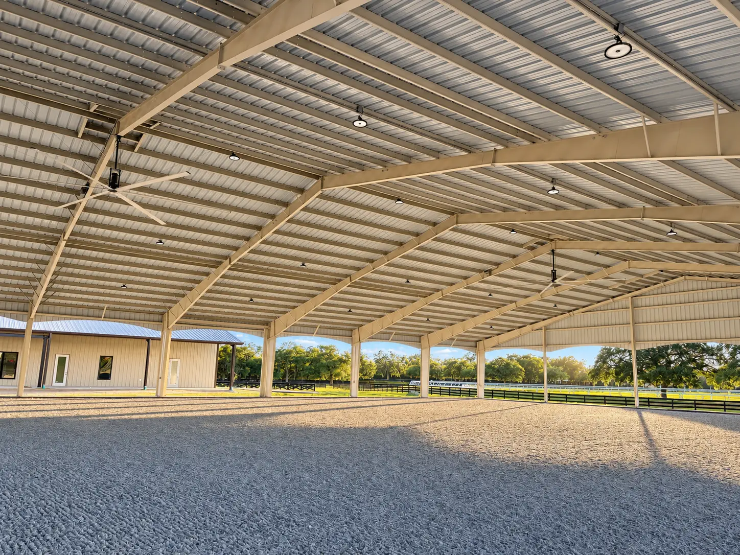 Inside the covered arena — sand footing beneath steel trusses with lights and fans overhead, the office complex beyond