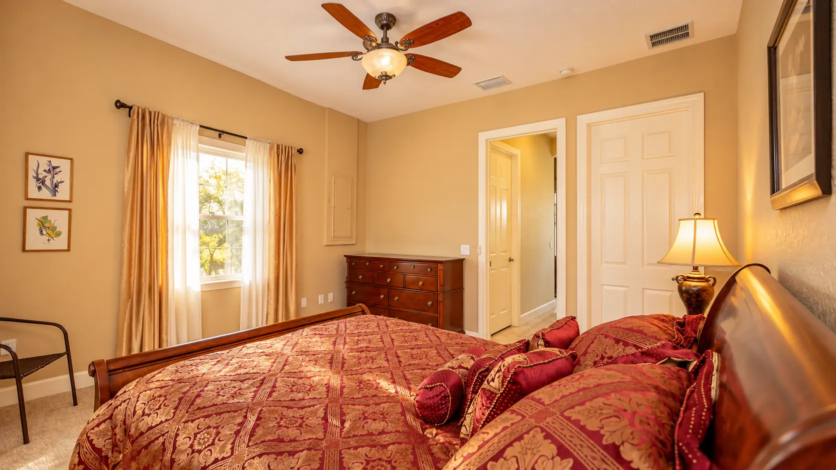 A bedroom in the viewing apartment with a wooden sleigh bed in red and gold bedding, a nightstand lamp, dresser, ceiling fan and draped window