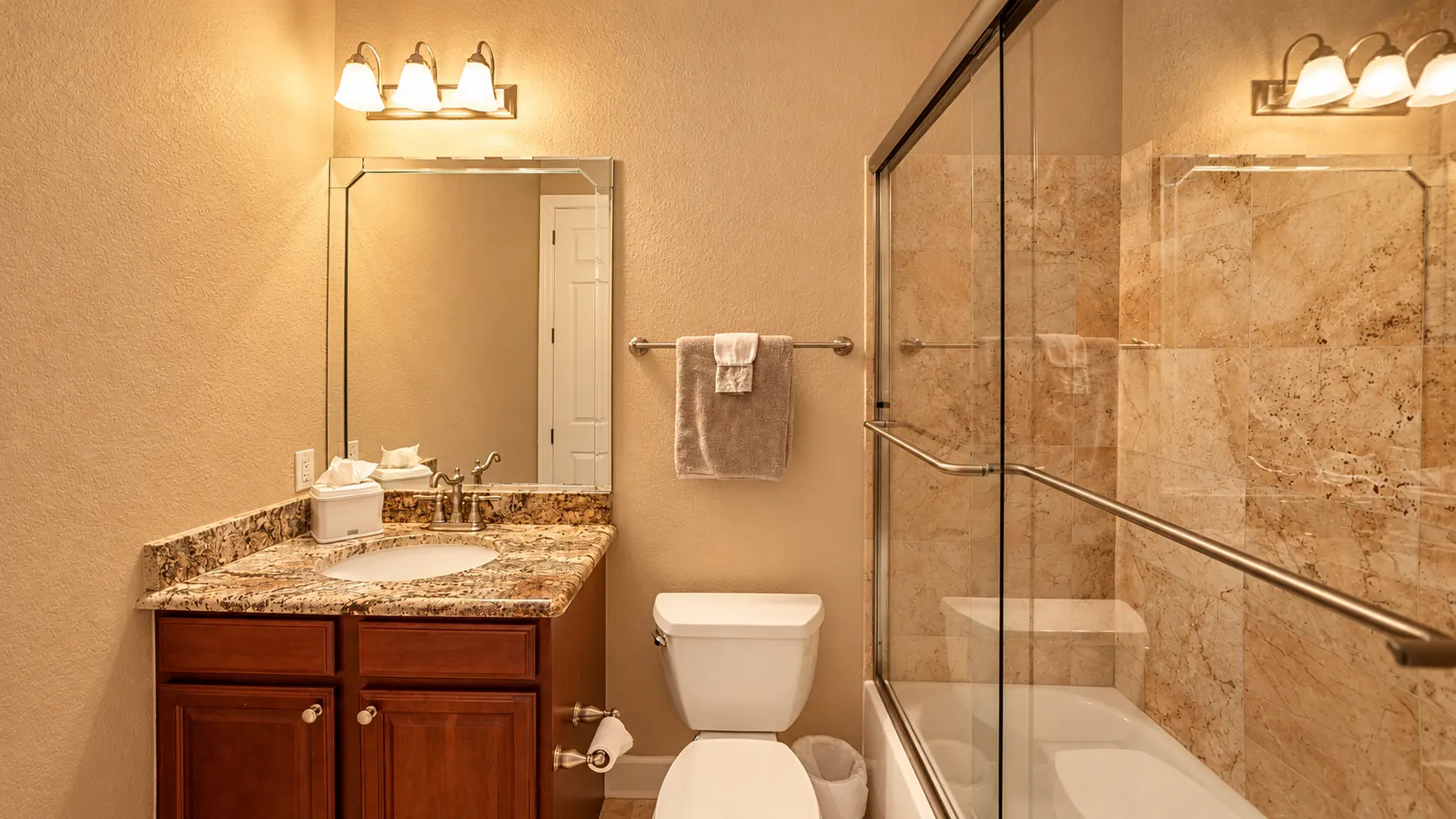 The apartment bathroom — a dark wood vanity with granite top beneath a mirrored light bar, beside a tub and glass shower