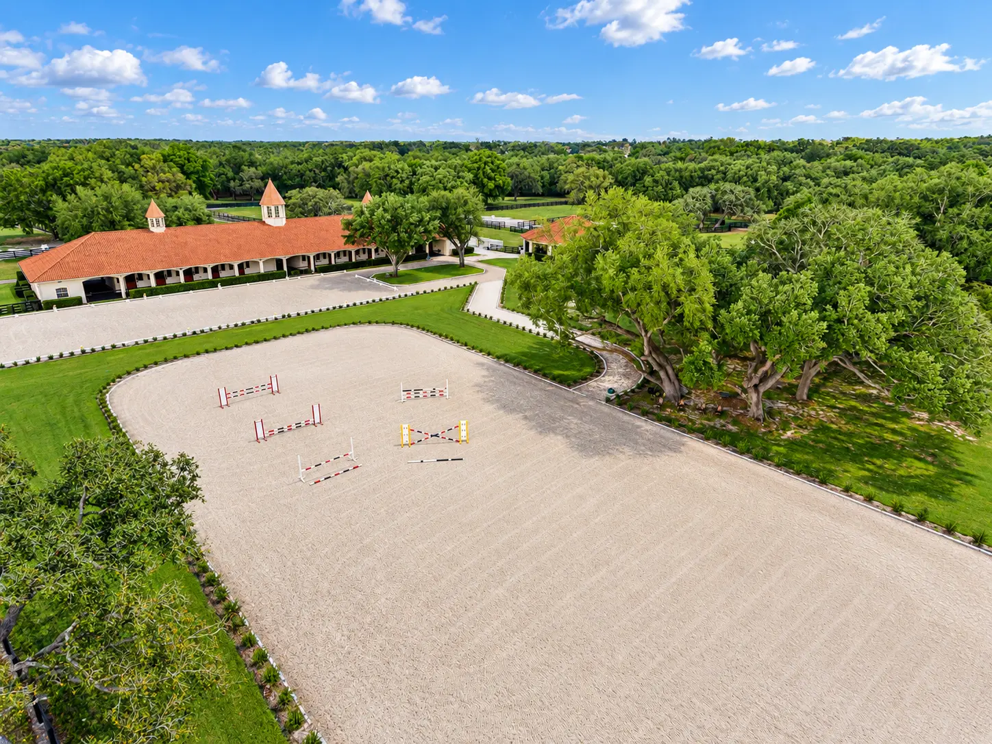 Aerial view of the sand jumping arena with jumps grouped at one end and the terracotta-roofed barn beyond